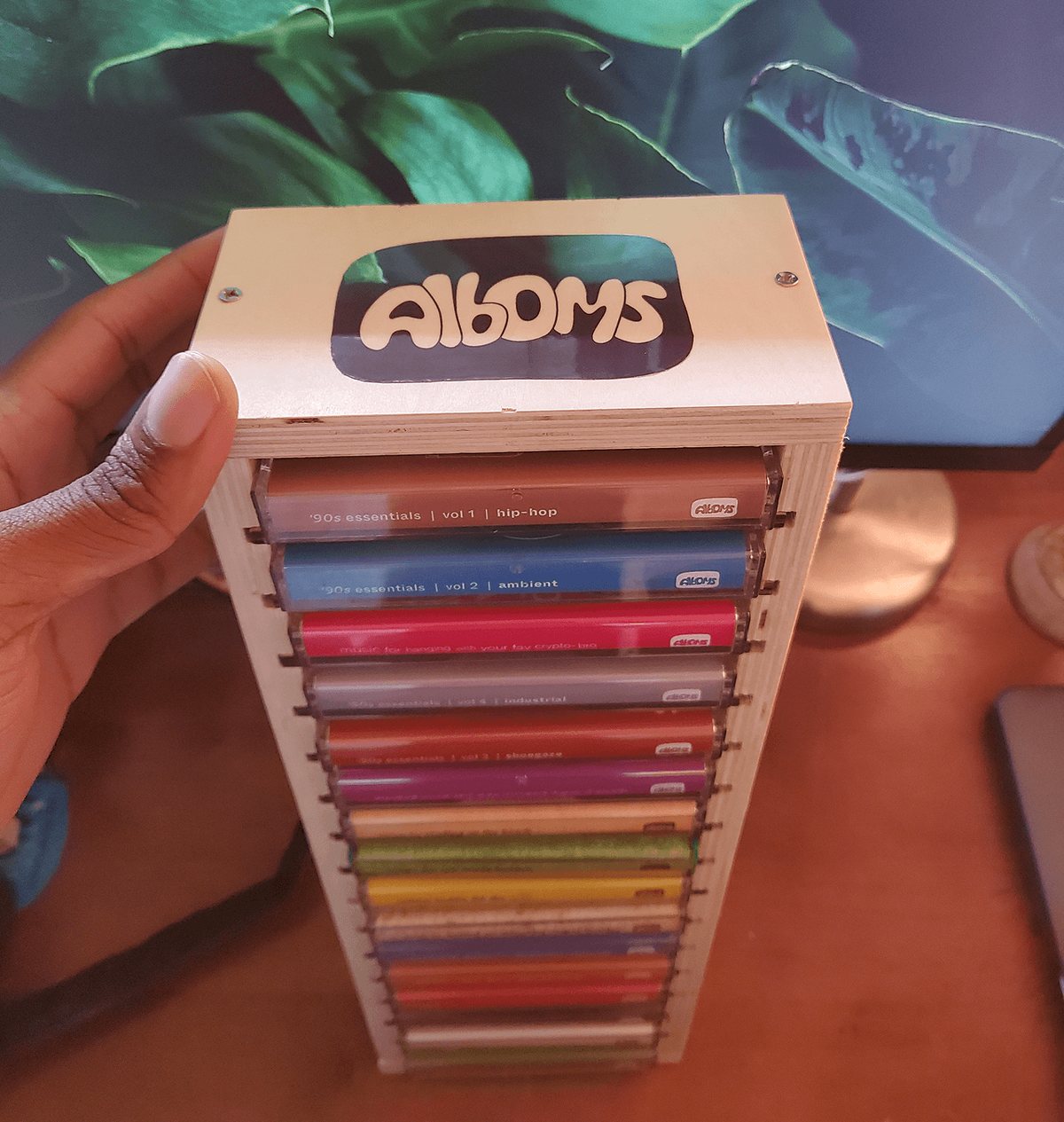 A hand holding a pale wooden Alboms rack, the bubble-letter badge cut out of its top, stacked with a rainbow of J-card spines