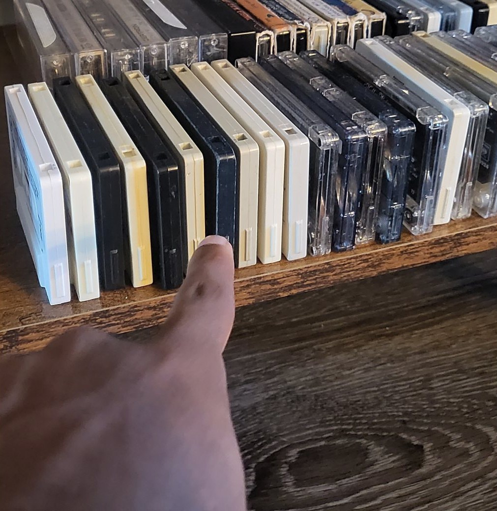 A finger pointing along a shelf of plain black, white, and clear cassette cases, none of them labeled